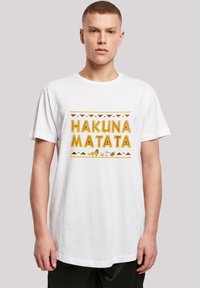 Bílé bavlněné tričko s nápisem "HAKUNA MATATA" tištěným tučnými žlutými písmeny, s grafickými obrázky zvířat a geometrickým vzorem v oranžové a hnědé barvě.
