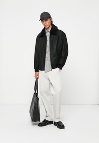 Black leather bomber jacket with a shearling collar, gray plaid shirt, white trousers, black shoes, and a gray tote bag. Minimalist design.