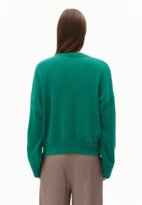 Person mit langen braunen Haaren, der einen lockeren grünen Pullover und beige Hosen trägt, von hinten betrachtet vor einem schlichten weißen Hintergrund.