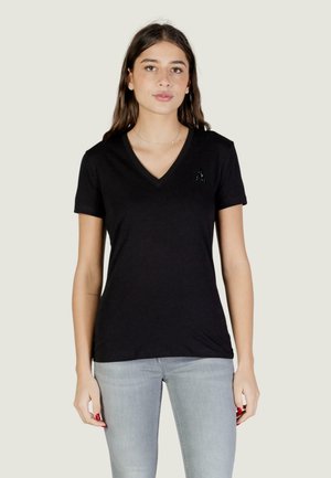 Giovane donna che indossa una T-shirt nera con scollo a V e un piccolo logo "A" sul petto, e jeans grigio chiaro, in piedi davanti a uno sfondo semplice.