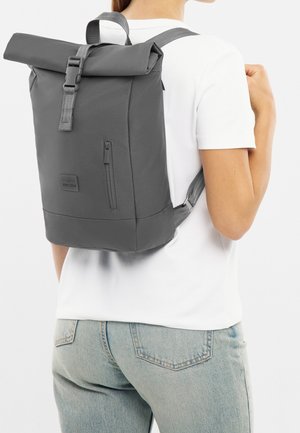 Šedý batoh s rolovacím zavíráním, nastavitelnými popruhy, boční kapsou na zip a texturou látky. Minimalistický design a detail loga na přední straně.