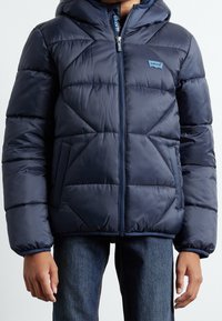 Veste matelassée bleu marine avec capuche, présentant un dessin matelassé en losanges, une fermeture éclair à l'avant, et le logo Levi's sur la poitrine. Tissu synthétique texturé.