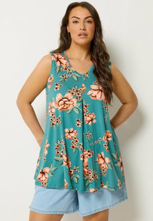 Kvinde med langt hår iført en ærmeløs teal top med orange blomsterprint og lyseblå denimshorts, stående med hænderne på hofterne.