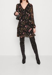 Nainen, joka on pukeutunut mustaan kukalliseen mekkoon, jossa on läpinäkyvät pitkät hihat, mustiin polven yli ulottuviin saappaisiin, ja pitää kädessään pientä mustaa käsilaukkua valkoista taustaa vasten.