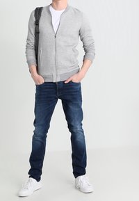 Grå herringbone zip-up jacka, vit T-shirt, mörkblå jeans och vita sneakers. Jackan har ribbade mudd och nederkant.
