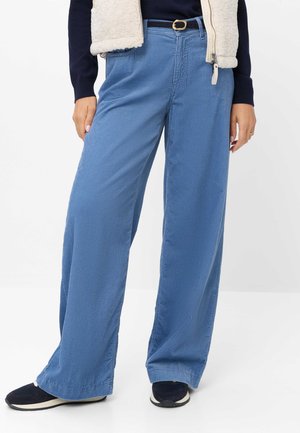 Personne portant un pantalon large en velours côtelé bleu avec une ceinture noire, des baskets noires, un haut bleu marine et une veste polaire beige.