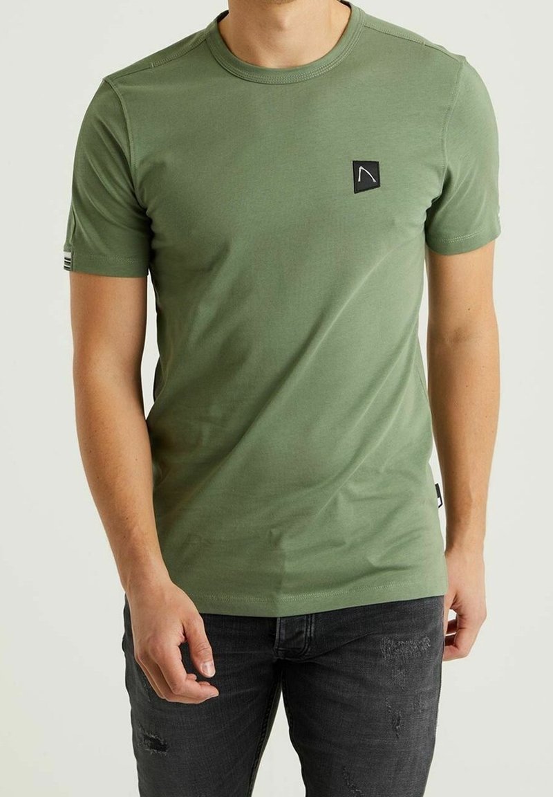 T-Shirt mit kurzen Ärmeln in Olivgrün aus Baumwolle. Mit einem schwarzen Logo-Patch auf der Brust und kontrastierenden Nähten. Kombiniert mit dunklen Jeans.