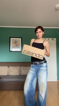 Hnedá krabica Zalando s tučným čiernym písmom, ktorú drží osoba v čiernom tielku a svetlomodrých širokých džínsach, v zelenej miestnosti.