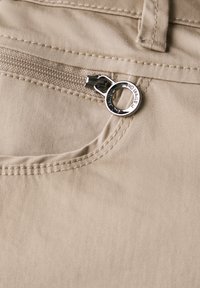 Primer plano de una tela beige que presenta un bolsillo con cremallera y un tirador plateado, costuras visibles y una textura suave.