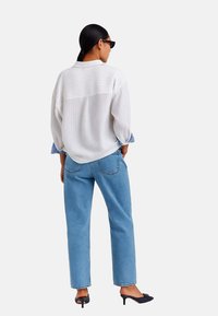 Maglietta bianca a coste oversize con polsini a righe blu, abbinata a jeans dritti azzurri e mule nere con tacco, vista da dietro.