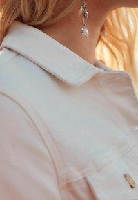 Chaqueta de mezclilla de color claro con un cuello clásico y botones frontales. Presenta una tela texturizada y una vista en primer plano de un pendiente con detalles de conchas marinas.