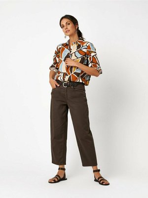 Woman standing with one hand in pocket wearing abstract patterned shirt, dark cropped pants, black belt, and black sandals.