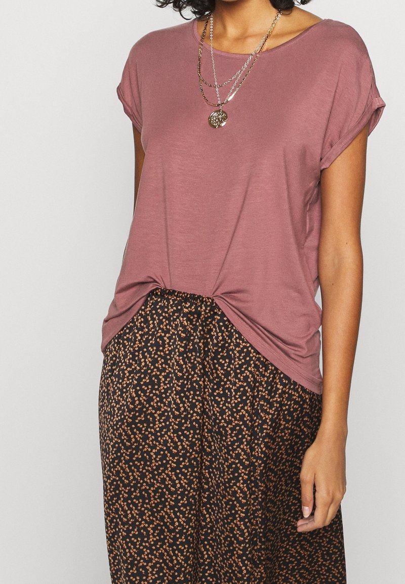 Korte mouw mauve t-shirt met een knoopdetail aan de onderrand, gecombineerd met zwarte broek met een oranje bloemmotief. Zachte stoftextuur.