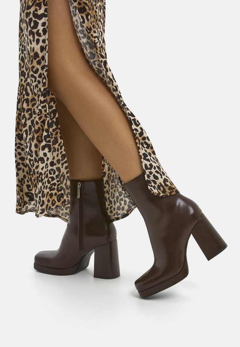 Femme portant des bottines en cuir marron à talons hauts et une jupe à imprimé léopard avec une fente haute, debout sur un sol blanc.