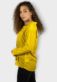 Camisa amarilla de manga larga con cuello abotonado y bolsillo frontal. Textura suave, corte relajado y hem redondeado. Combinada con pantalones negros.