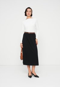 Λευκή μπλούζα με μακριά μανίκια συνδυασμένη με μαύρη midi φούστα, σφιγμένη με καφέ ζώνη· το μοντέλο κρατάει καφέ τσάντα και φοράει μαύρα μυτερά τακούνια.