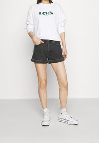 Sudadera blanca con logo verde de Levi's, combinada con pantalones cortos de mezclilla negros con puños enrollados y zapatillas blancas de caña alta. Diseño sencillo.