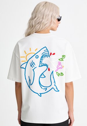 Personne aux cheveux blonds clairs portant un T-shirt blanc oversize avec un motif coloré de requin et de soleil dessin animé dans le dos, vue de dos.