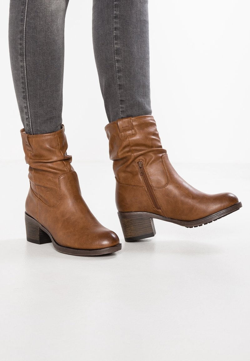 Bottines en cuir marron avec un design froncé, une fermeture éclair latérale, un bout arrondi et un talon épais en bois, portées avec un jean gris slim.