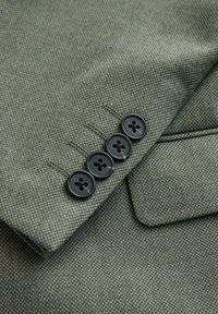 Nahaufnahme eines grünen, strukturierten Jackenärmel mit vier schwarzen Knöpfen und einer Klappentasche am Körper der Jacke.