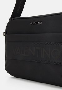 Černá nylonová taška přes rameno s texturovaným povrchem, vybavená vyraženým logem „VALENTINO“, nastavitelným popruhem a elegantním designem.