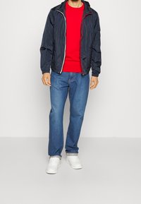 Marine let jakke med hætte, sølv lynlås, rød t-shirt med rund hals, blå jeans og hvide sneakers.