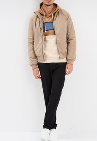 Giacca bomber beige sopra una felpa color block tan e blu, abbinata a pantaloni neri e scarpe da ginnastica bianche, in piedi contro uno sfondo bianco.