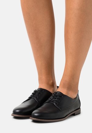 Chaussures en cuir noir avec une texture lisse, bout rond et un design à lacets. La fine semelle marron complète ce modèle épuré.