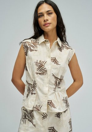 Mujer con blusa sin mangas color crema abotonada y pantalones cortos a juego con estampado de hojas marrones, de pie frente a un fondo gris liso.