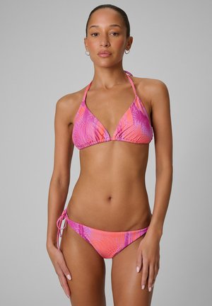 Kobieta w różowo-pomarańczowym wzorzystym bikini na szyję z wiązaniami po bokach, stojąca na prostym szarym tle, zwrócona przodem.