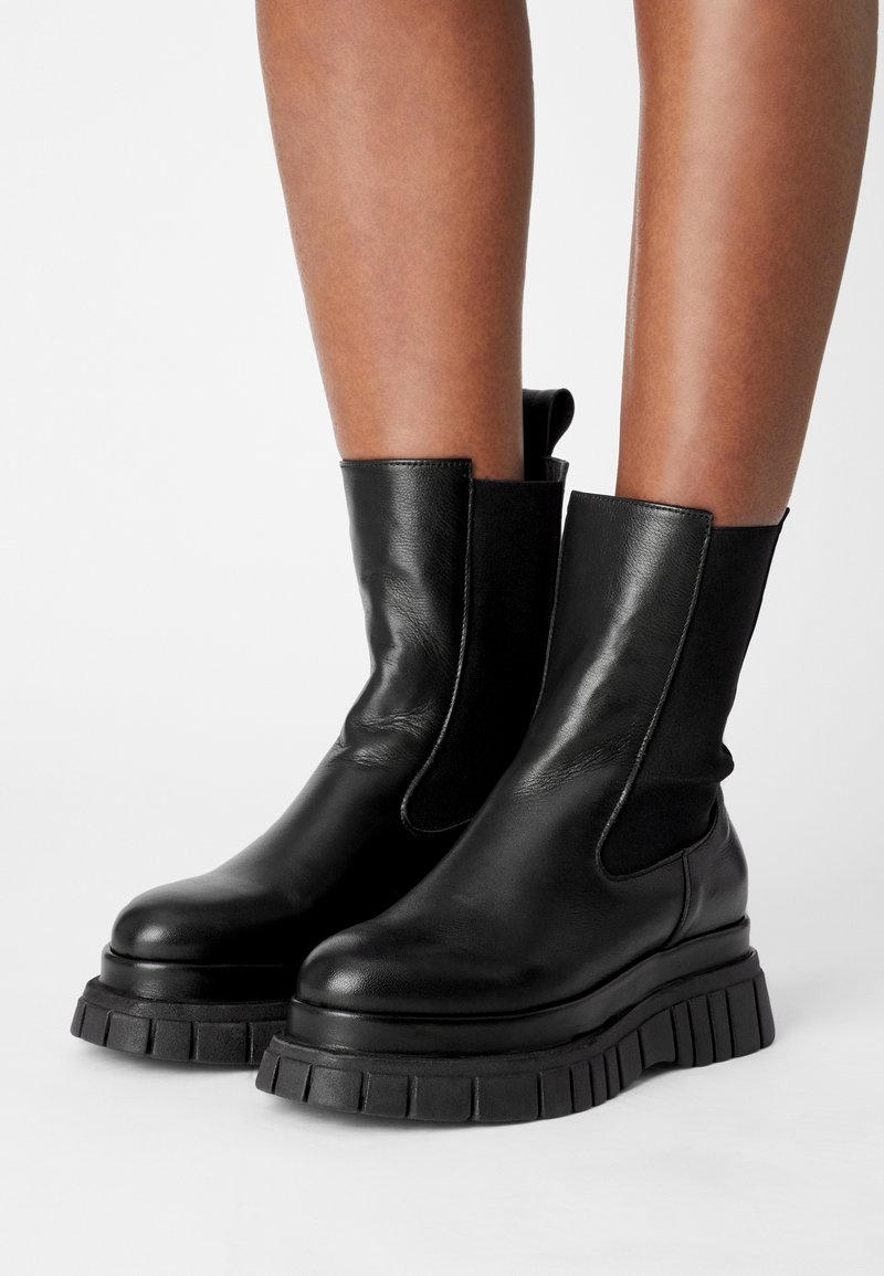 Jambes portant des bottines en cuir noir avec des semelles épaisses et crantées ainsi que des panneaux latéraux élastiques, sur un fond blanc uni.