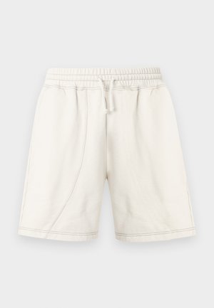 Mees, kes kannab tumesinist nokamütsi ja logoga t-särki, kreemikaid paelaga lühemaid pükse, musti sokke ja musti tossusid, seisab käed taskutes.