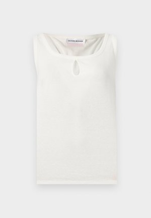 Vrouw draagt mouwloze crème top met sleutelgathals, hoge taille wijde donkere jeans, zwarte riem en zwarte open-teenhakken, staand.