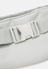 Ljust grå tygväska med en texturerad yta, en tunn rem och en plastspänne med geometrisk design.