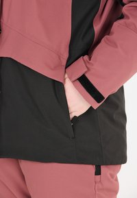 Rosa och svart jacka med en strukturerad yta, utrustad med en sidoficka och justerbara ärmslut med kardborreband.