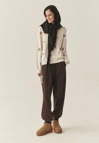 Kötött pulóver színes virág- és figura hímzéssel, laza barna nadrággal és bézs bokacsizmával párosítva. Fekete sál a nyak köré kötve.