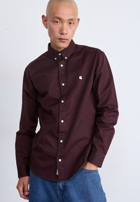 Camisa de botón de manga larga burdeos, hecha de algodón, con botones blancos y un pequeño logotipo en el lado izquierdo del pecho, combinada con pantalones de mezclilla.