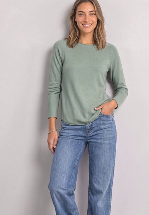 Jeune femme souriante, portant une chemise à manches longues vert clair et un jean bleu, posant avec une main dans la poche devant un fond uni.