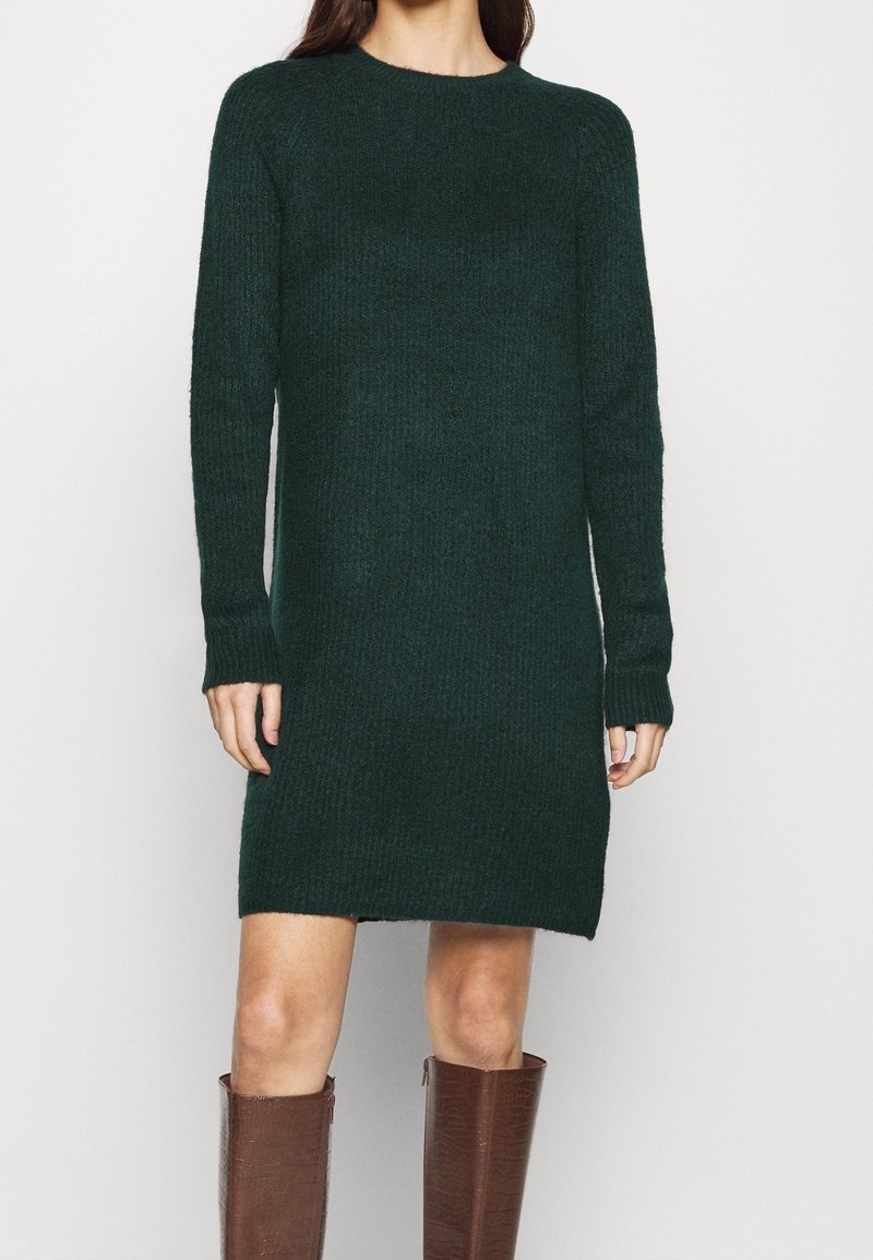 Robe tricotée à manches longues de couleur vert foncé. Présente un col rond, une coupe décontractée et une texture côtelée. Portée avec des bottes marron jusqu'aux genoux.