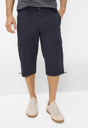 Mand iført marineblå cargoshorts til knæet og beige sneakers, stående med hænderne i lommerne mod en hvid baggrund.