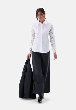 White shirt with lace long sleeves, paired with dark wide-leg trousers. Model holds a dark jacket, standing on a plain background.