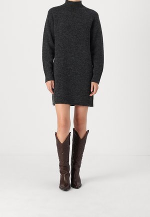 Personne portant une robe-pull tricotée à manches longues gris foncé et des bottes en cuir marron foncé montantes jusqu'aux genoux, debout devant un fond uni.