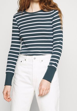 Femme portant un haut à manches longues à rayures horizontales vert foncé et blanc avec un jean blanc taille haute, vue des épaules aux hanches.