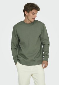 Sweatshirt vert à col rond en tissu doux, avec des manches longues, des poignets côtelés et une coupe décontractée, associé à un pantalon de couleur claire.
