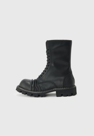 Μαύρο μποτάκι μάχης με κορδόνια, χοντρή σόλα με ανάγλυφα και λεπτομέρεια από μαλακό δέρμα στην περιοχή των δακτύλων, απεικονίζεται σε πλευρική όψη σε απλό φωτεινό φόντο.