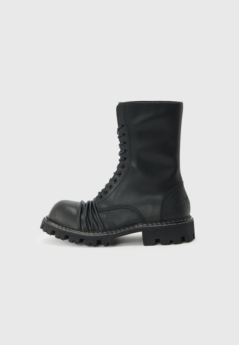 Μαύρο μποτάκι μάχης με κορδόνια, χοντρή σόλα με ανάγλυφα και λεπτομέρεια από μαλακό δέρμα στην περιοχή των δακτύλων, απεικονίζεται σε πλευρική όψη σε απλό φωτεινό φόντο.