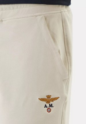 Tasca di pantaloni beige con emblema ricamato di un'aquila dorata, corona, iniziali "A.M." e un piccolo emblema circolare in rosso, bianco e verde.
