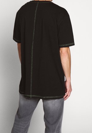 Mann trägt ein schwarzes kurzärmeliges Hemd mit kontrastreichen weißen Nähten entlang des Rückens und an den Saumkanten, kombiniert mit grauer Hose.