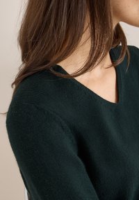 Grüner Pullover mit V-Ausschnitt, aus weichem Stoff gefertigt, mit gerippten Details an den Schultern. Nahaufnahme von Textur und Ausschnitt.