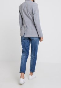 Blazer a quadro in grigio chiaro e grigio scuro, con uno spacco singolo e una forma su misura, abbinato a jeans blu sbiaditi e sneakers bianche.